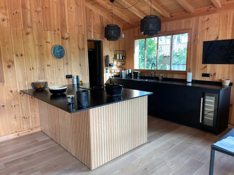 Cuisine sur mesure dans une maison à ossature bois : îlot central à façade cannelée en chêne, plan de travail noir et meubles bas laqués noirs